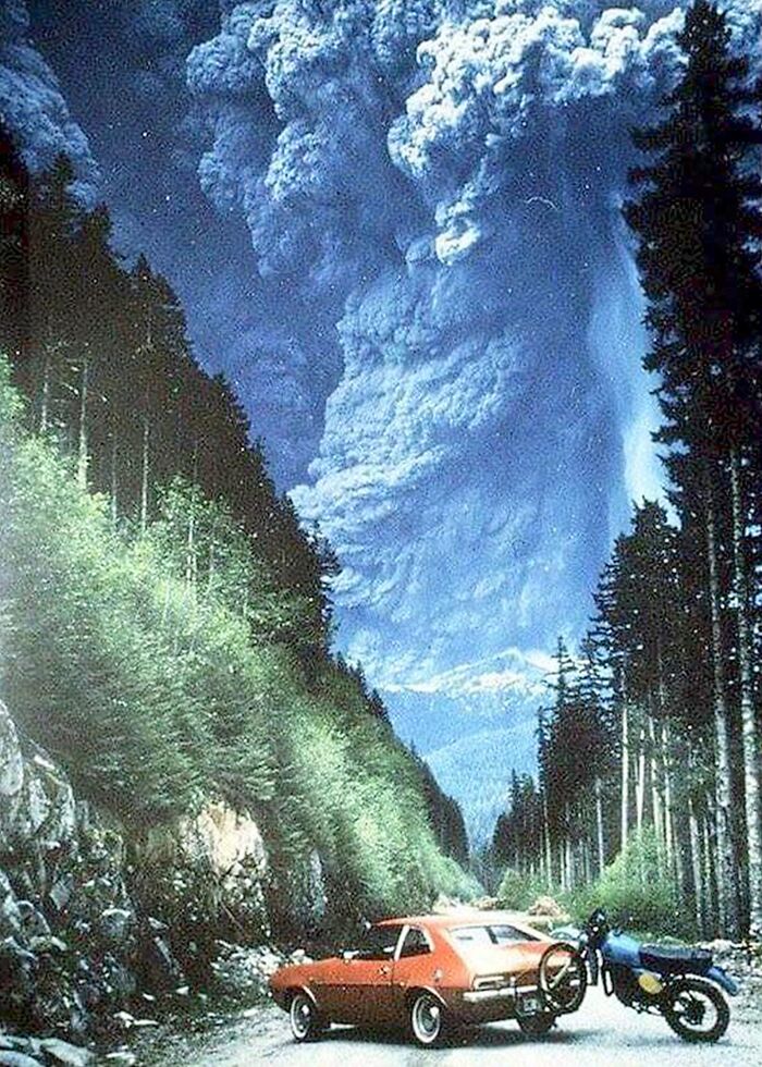 Red car and motorcycle parked on a forest road with a massive volcanic eruption cloud in the background, 1980s photo.
