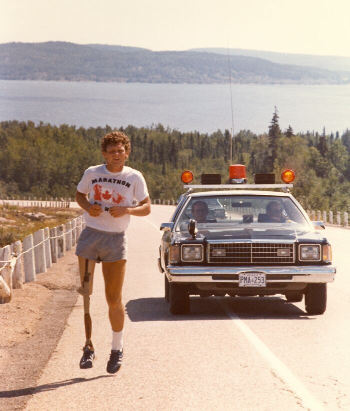 Runner with a prosthetic leg jogging on a scenic road alongside a police car in a wild and wonderful 1980s setting.