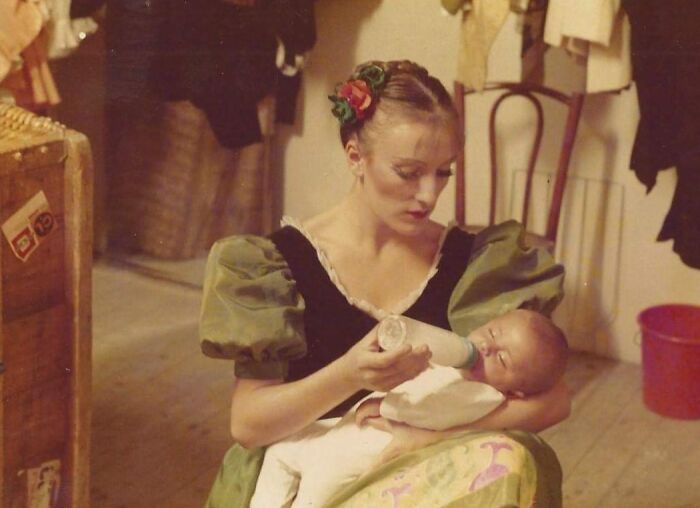 Woman in 1980s dress feeding baby with a bottle inside a vintage room with wooden furniture and clothing.