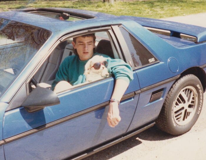 Young man with dog wearing sunglasses in a blue sports car, showcasing wild and wonderful 1980s style.
