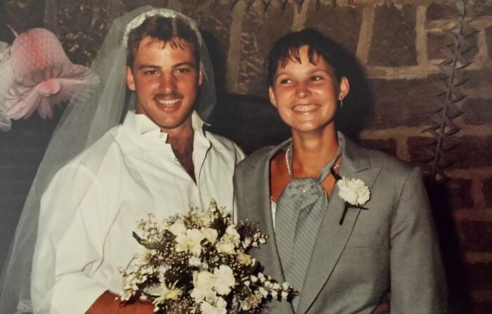 Couple dressed in quirky 1980s wedding attire smiling and holding a bouquet in a rustic setting.