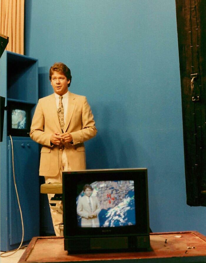 1980s weather forecast studio with man in beige suit and vintage TV showing weather map in a wild and wonderful setting