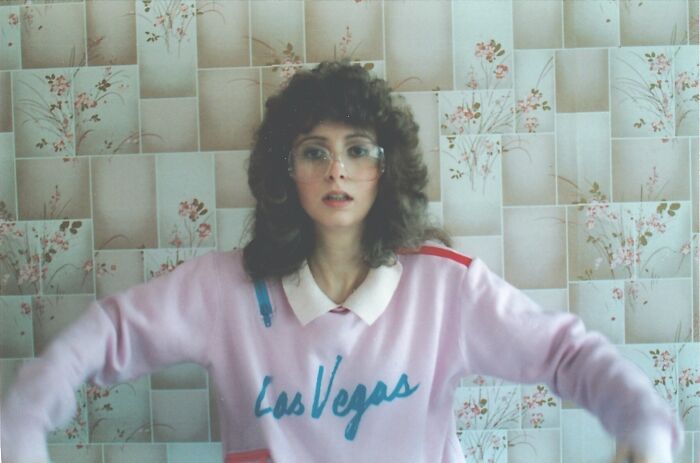 Woman with curly hair and large glasses wearing a pink Las Vegas sweater, showcasing wild and weird 1980s fashion style.