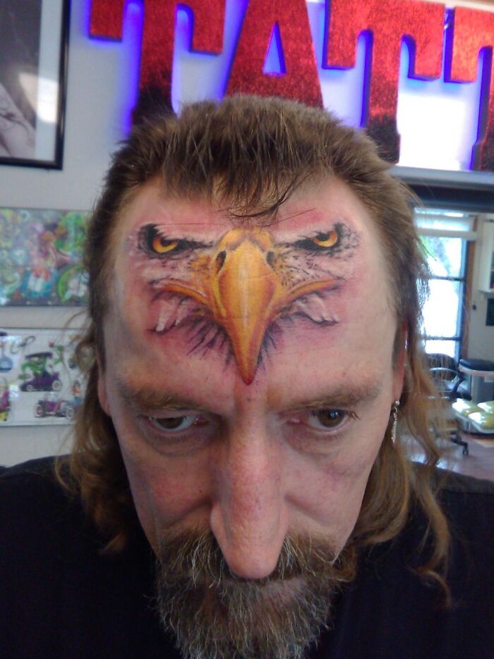 Man with a tattoo fail of an eagle on his forehead blending oddly with his facial features in a tattoo studio.