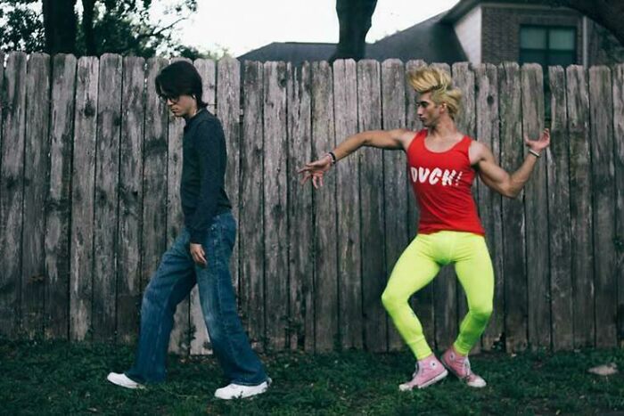 Two people in a yard, one dressed casually and the other wearing bright clothes and striking a humorous pose, people chuckling.