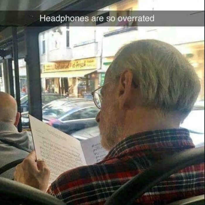 Older man reading sheet music on a bus while a sign reads headphones are overrated in a humorous setting.