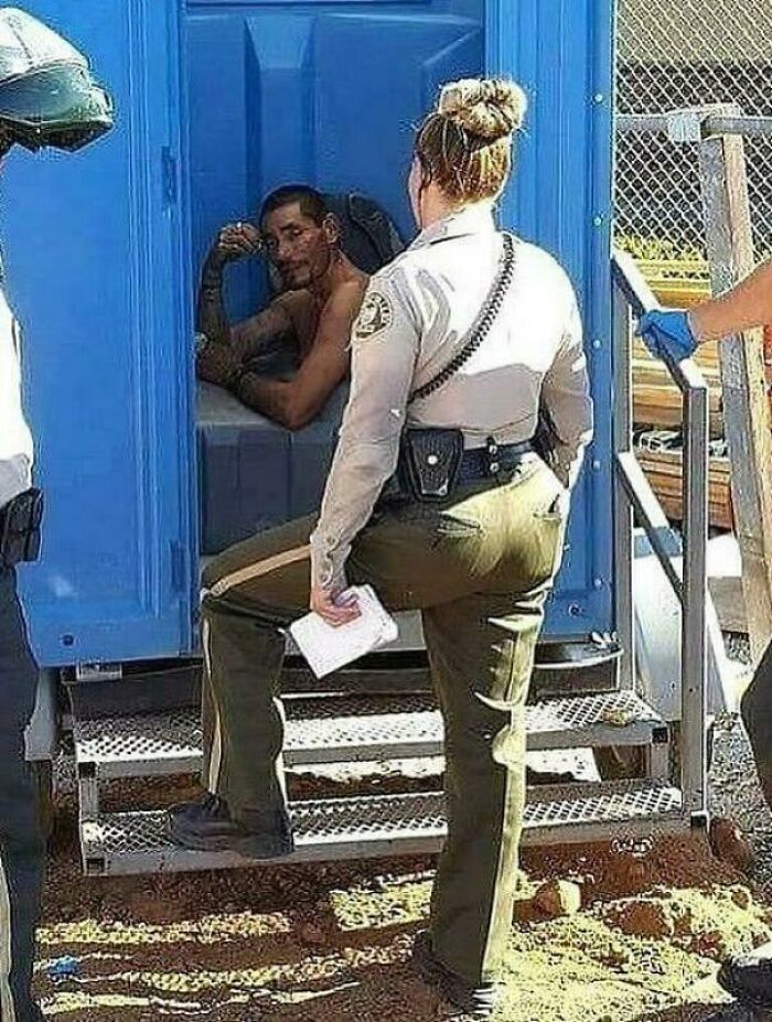 Man sitting inside a portable toilet talking to a female officer outside, a humorous moment people are chuckling at.