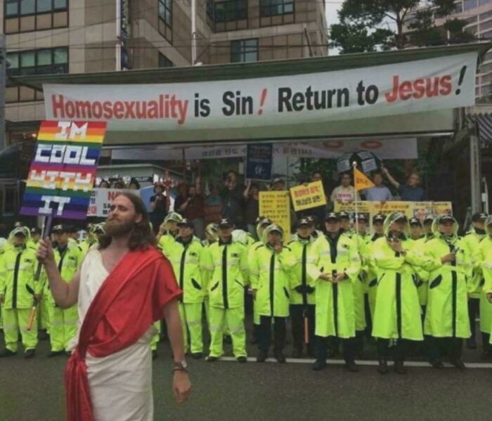 Person dressed as Jesus holding a pride flag with police and protestors behind in a picture that goes hard.