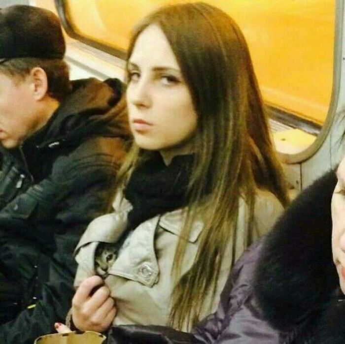 Young woman on public transit with a small cat hidden inside her coat, a picture that goes hard and makes people chuckle.
