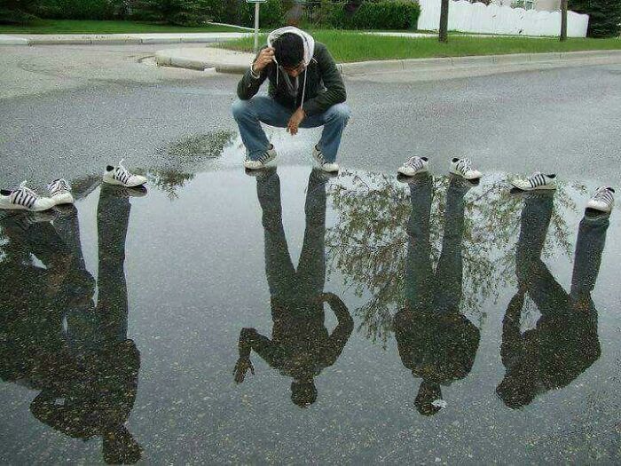 Man squatting by a puddle with reflections showing multiple people without shoes in a humorous picture that goes hard.