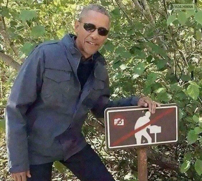 Man wearing sunglasses posing next to a no photography sign in a wooded area, a humorous picture that goes hard.