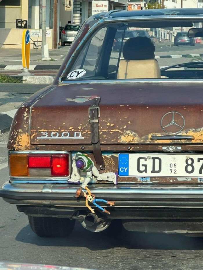 Rusty classic Mercedes 300D with Toy Story figures Buzz and Woody hanging from the rear bumper, people chuckling at this picture.