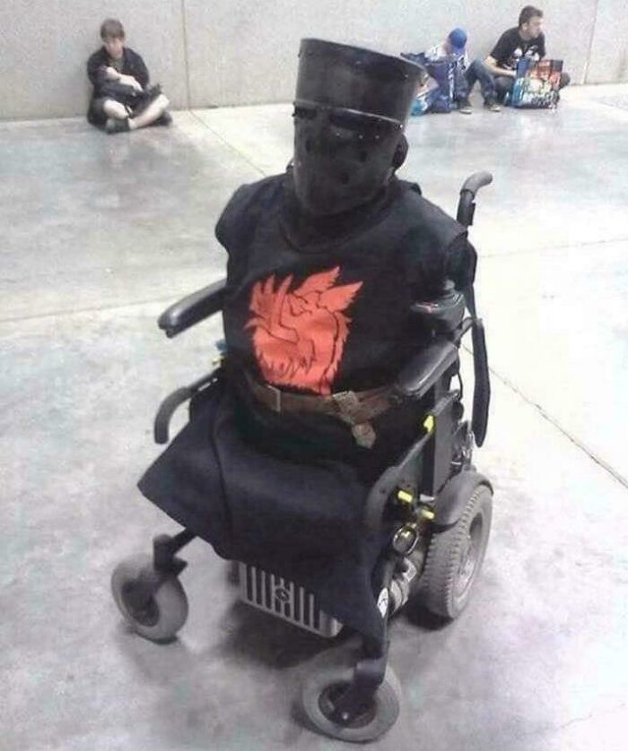 Person in an armored helmet and dark clothing sitting in a motorized wheelchair, a photo that goes hard and makes people chuckle.