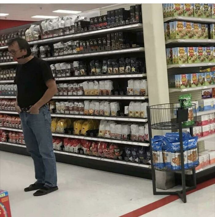 Man wearing a gag mask standing in Walmart aisle surrounded by shelves of packaged snacks and groceries.