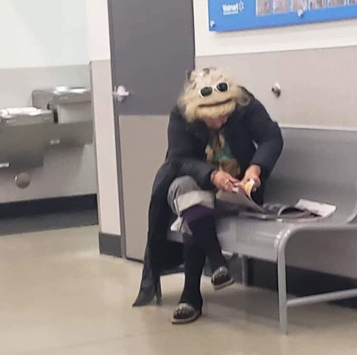 A person dressed unusually sitting on a bench reading a magazine in a public indoor setting, people of Walmart.
