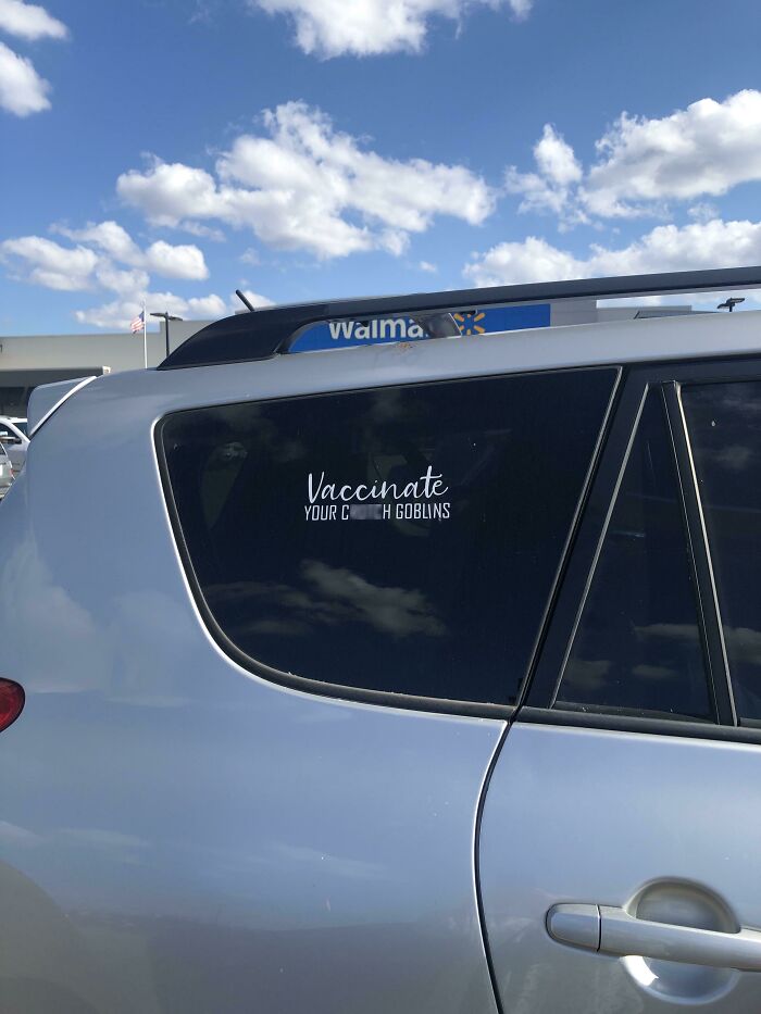 Sticker on a car window in a Walmart parking lot reading vaccinate your goblins, captured in a bright sky setting.