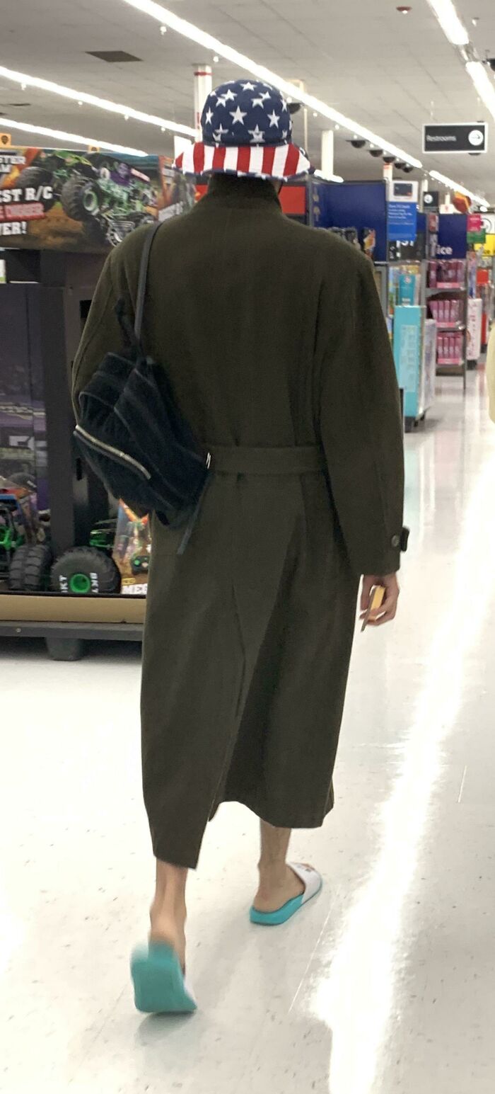 Person wearing a patriotic star-spangled hat, long coat, and slippers walking in Walmart aisle near toys section.