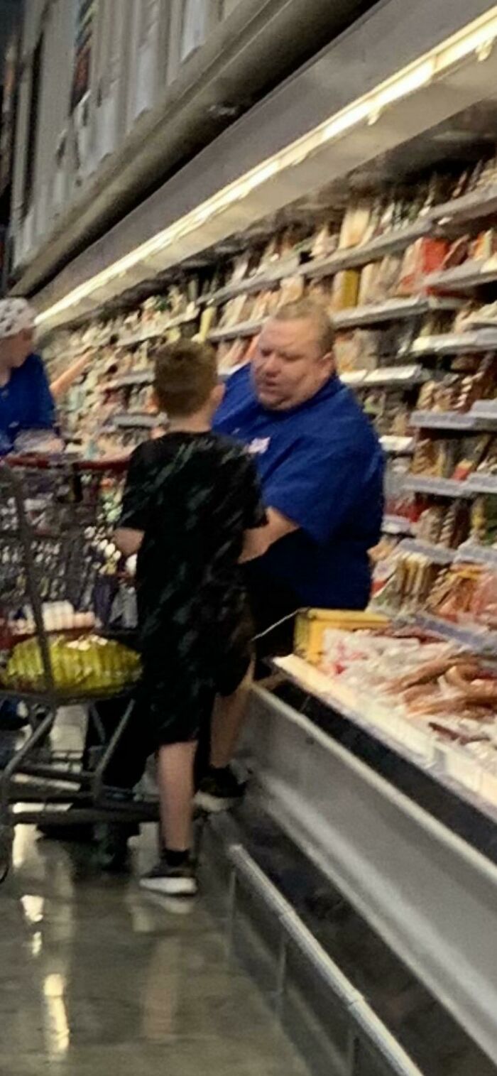 Man and child in Walmart deli aisle, surrounded by stocked shelves and a shopping cart with groceries inside.