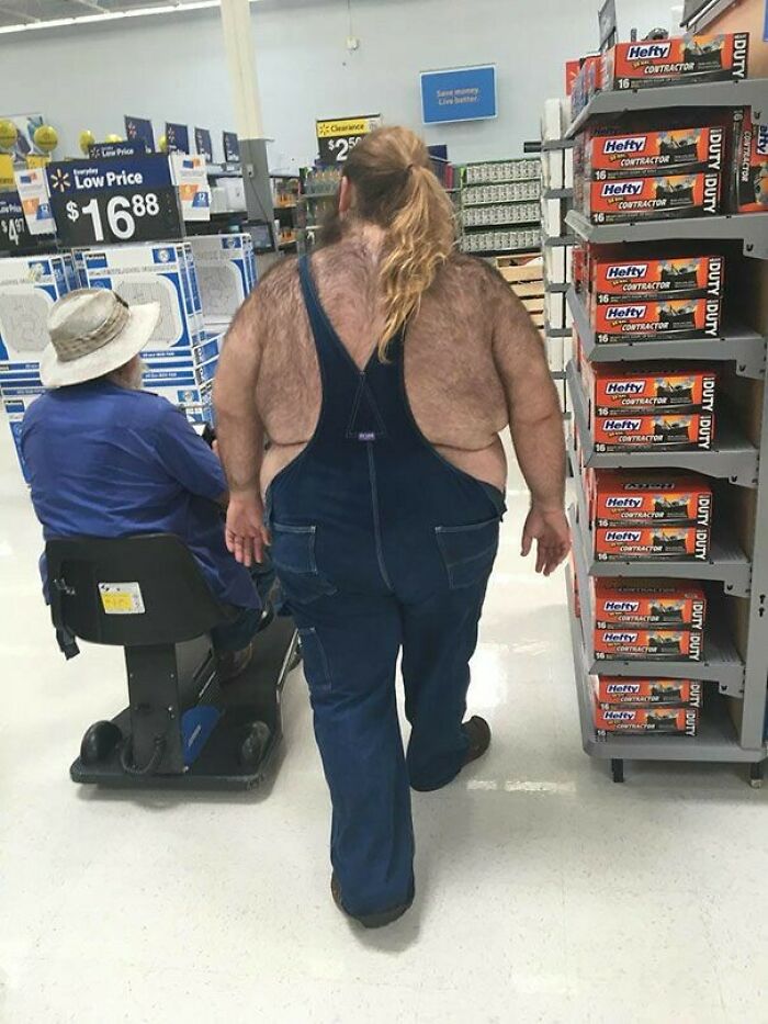 Person with visible back hair wearing overalls walking past a man on a mobility scooter in a Walmart aisle.