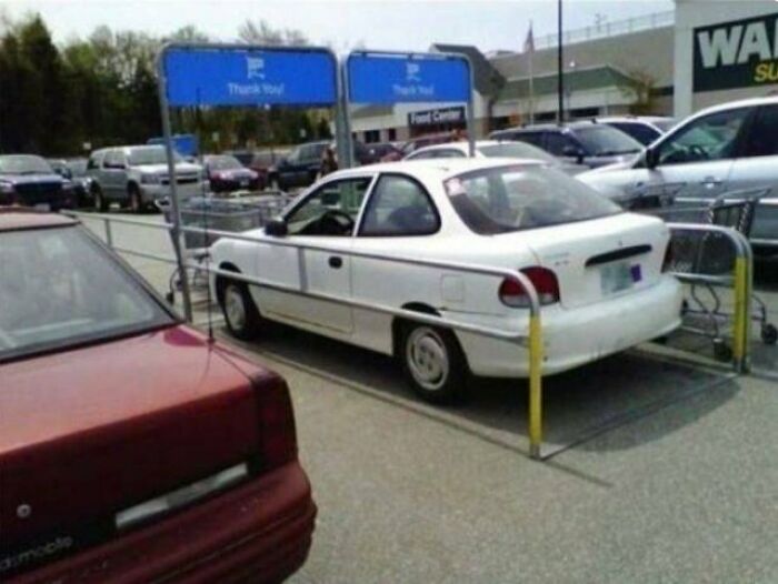 White car parked improperly in a Walmart parking lot, surrounded by shopping cart barriers, unusual People of Walmart moment.