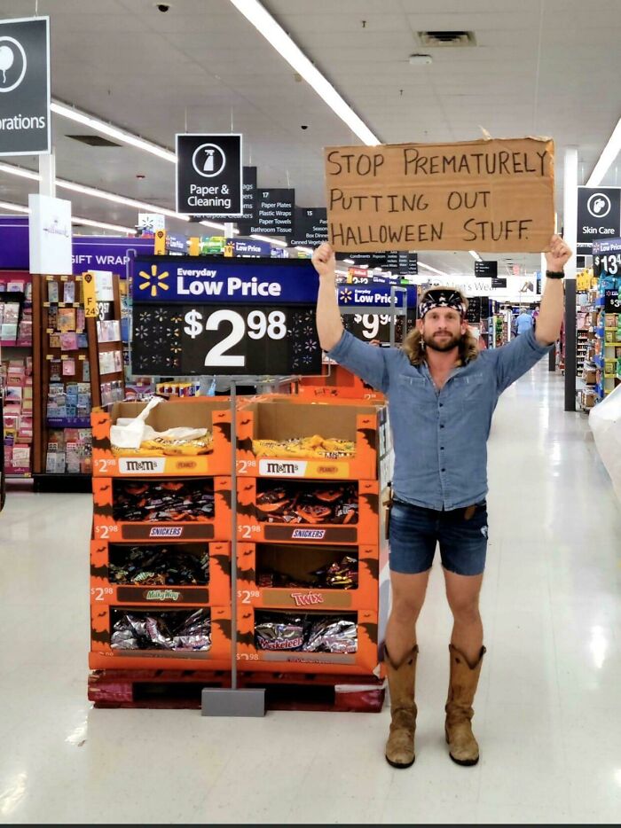 Man in cowboy boots and shorts holding a sign inside Walmart near Halloween candy display in a quirky People of Walmart moment
