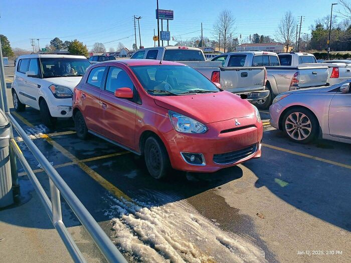 Red compact car parked in a crowded Walmart parking lot surrounded by various trucks and vehicles on a sunny day.
