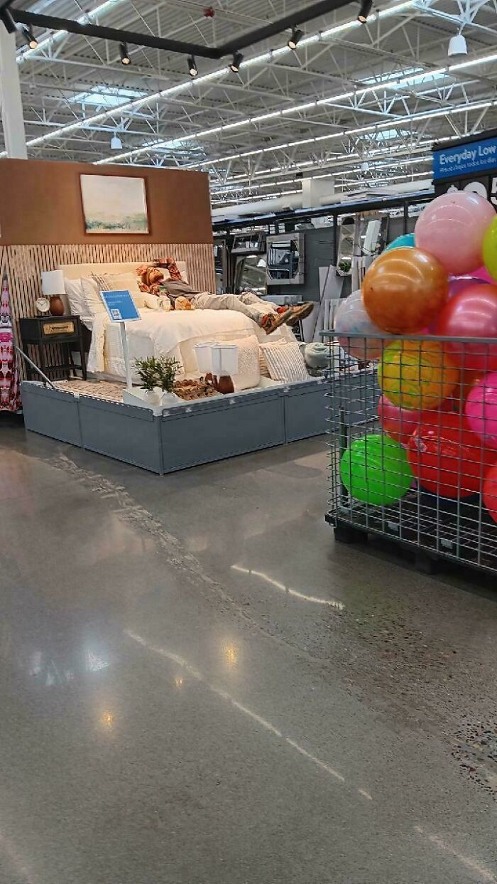 Person lying on a retail display bed inside Walmart store with colorful balls in a wire basket nearby.