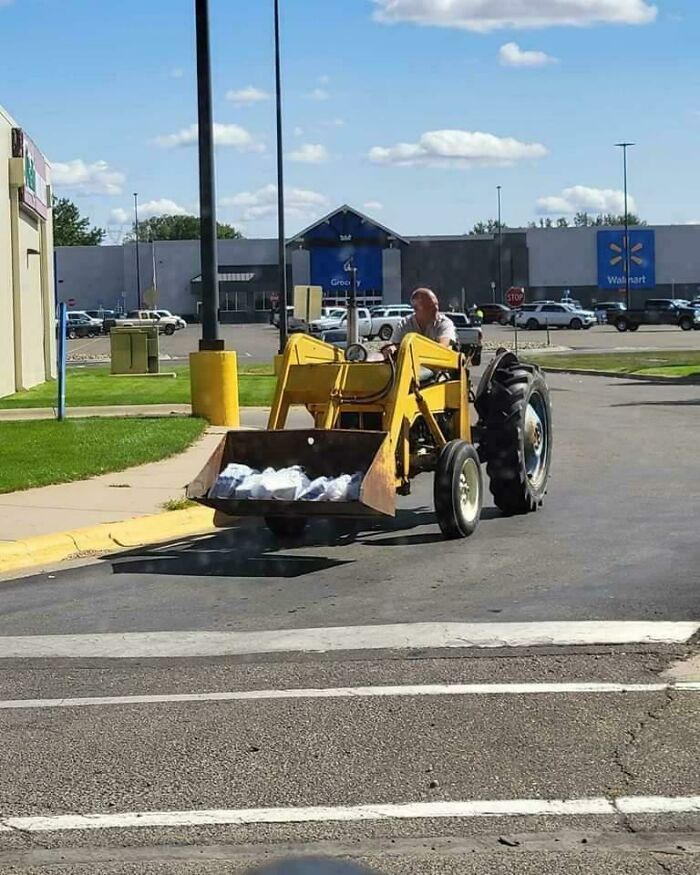 Man driving a yellow tractor in a Walmart parking lot with bags in the front loader on a sunny day.