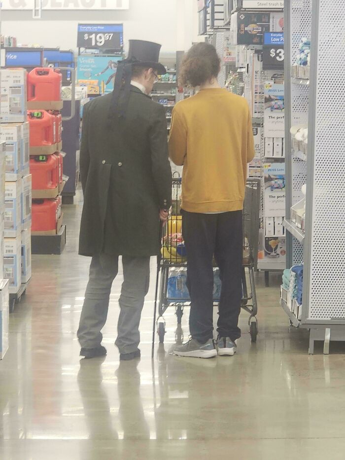 Two shoppers in a Walmart aisle, one wearing a vintage coat and top hat, showcasing unique People of Walmart style.