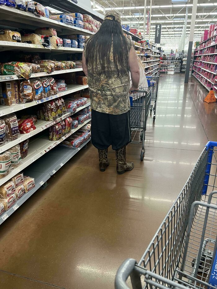 Man with long hair wearing camouflage and boots pushing a shopping cart in a Walmart aisle with bread products.