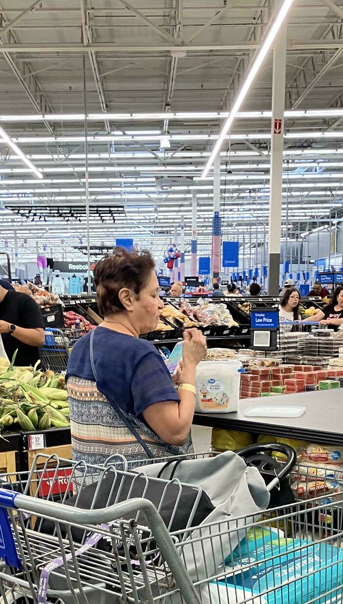 Woman eating at checkout counter inside Walmart with full shopping cart in busy store featuring produce and packaged goods