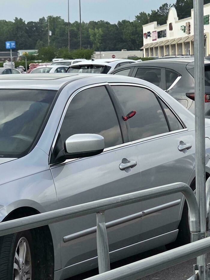 Silver car parked in Walmart parking lot with a small red toy hammer oddly attached to the side window.