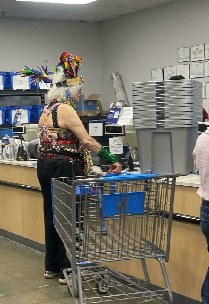 Eccentric person wearing a colorful feathered headpiece and unique outfit standing with a shopping cart inside Walmart.
