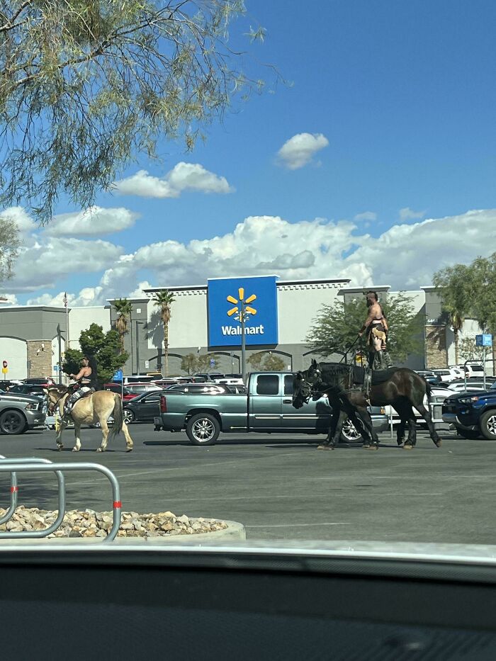 Two people riding horses in a Walmart parking lot under a clear blue sky with scattered clouds.