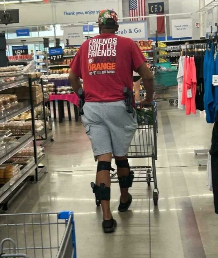 Man wearing knee holsters and shorts pushing a shopping cart inside a Walmart store aisle.