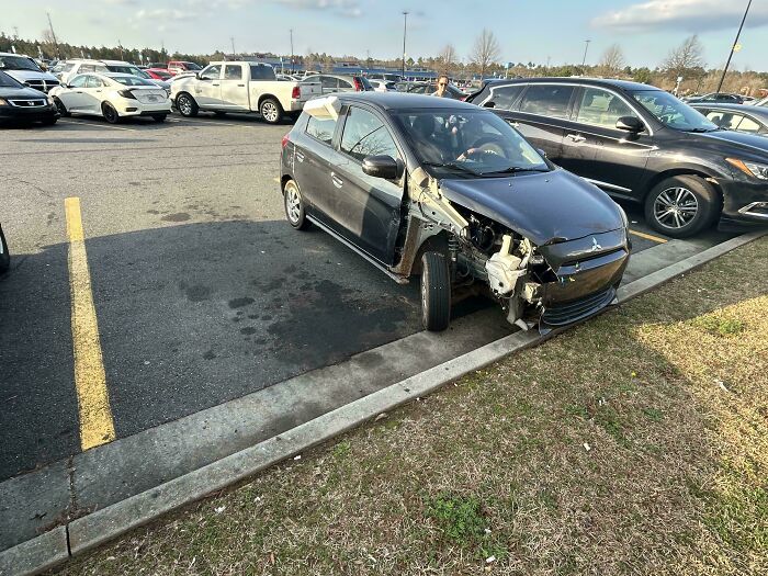 Damaged black car with front missing parts parked unevenly in busy Walmart parking lot during daytime