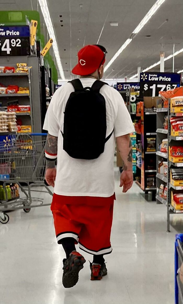 Man in red shorts and cap wearing a black backpack walking down an aisle inside Walmart store with snacks on shelves.