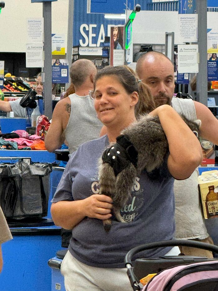 Woman holding a raccoon inside a Walmart store with other shoppers and checkout counters in the background.