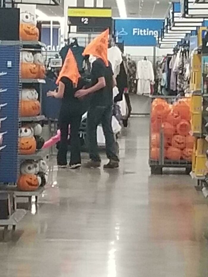 Two people wearing orange pumpkin hats shopping in a Walmart aisle filled with Halloween decorations.