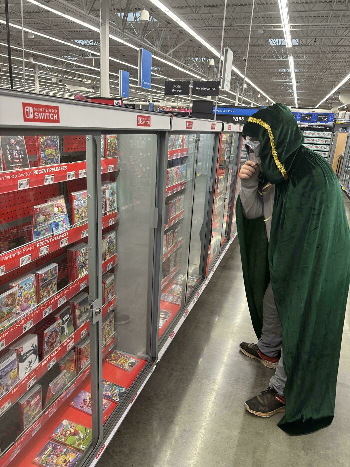 Person wearing a green hooded cloak and mask browsing Nintendo Switch games at Walmart’s video game aisle.