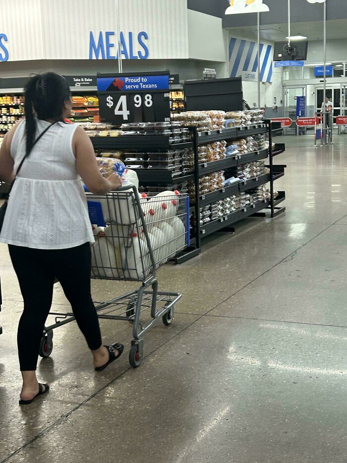 Woman pushing a shopping cart filled with goods inside a Walmart store aisle showing bakery and meal sections.
