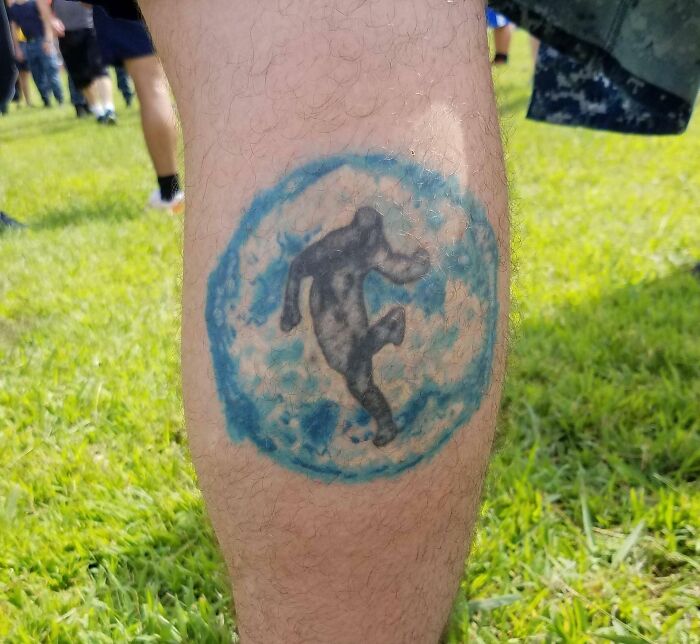 Tattoo fail on calf showing a blurry, poorly executed blue and black design with an indistinct figure inside a circle outdoors.