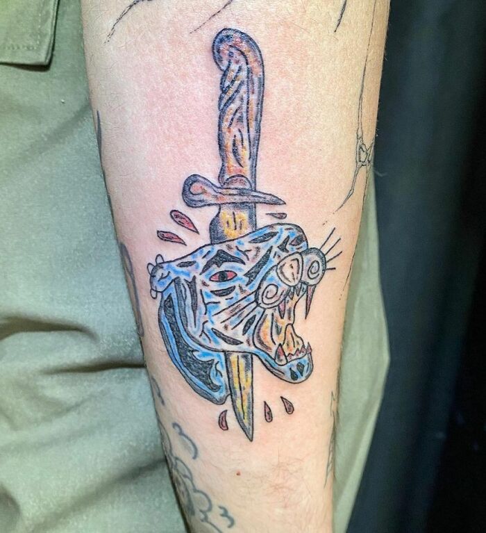 Tattoo fail showing a poorly executed blue panther head pierced by a dagger with blood drops on a forearm.