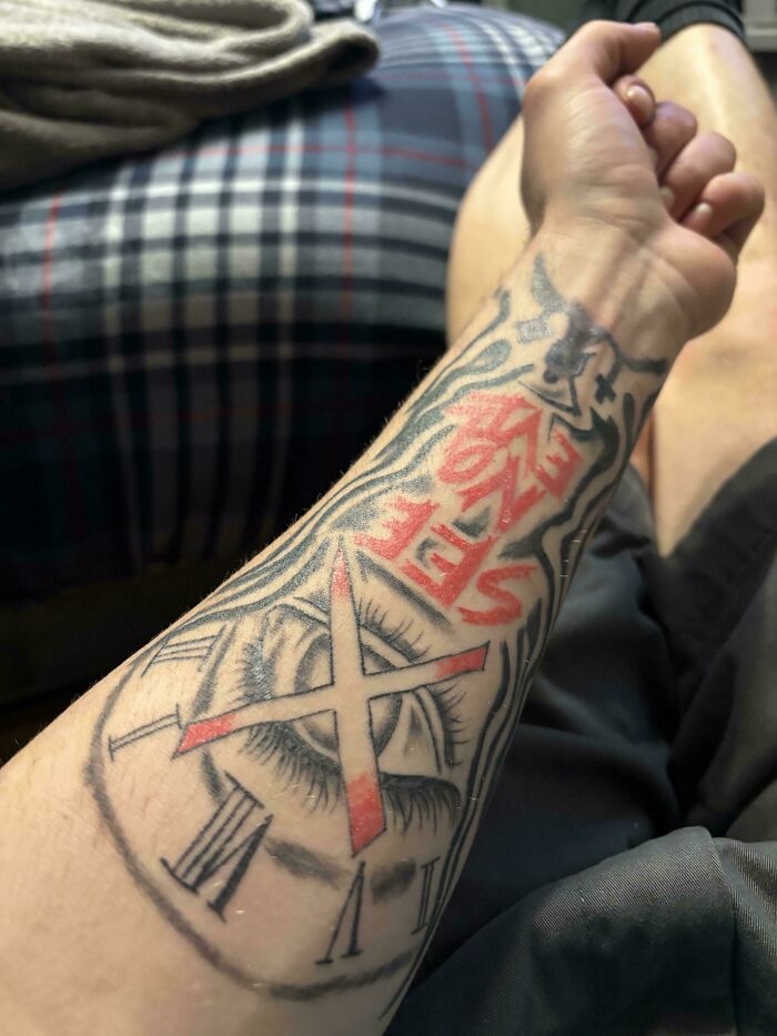 Forearm tattoo featuring an eye with clock numbers and bold red and black ink, showcasing a unique but flawed design.