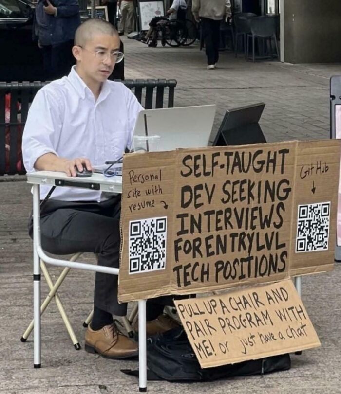 Man working on laptop outdoors with cardboard sign seeking self-taught developer interviews for entry level tech positions programming memes