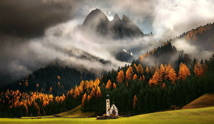 Autumn leaf colors and fall foliage glowing on a forested mountainside with mist and a small chapel below.