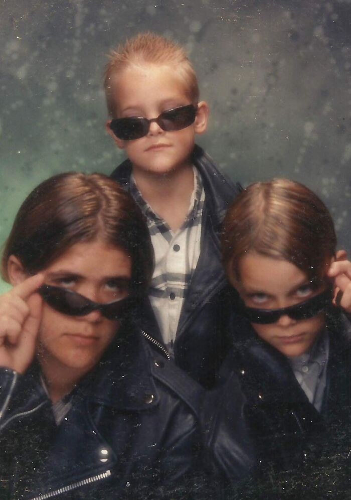 Tres hermanos con gafas oscuras y chaquetas de cuero posando para una foto familiar incómoda y divertida.