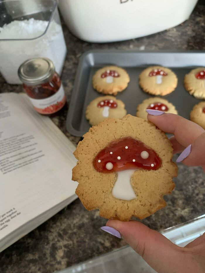 Hand holding a mushroom-decorated cookie with more on a tray in a cozy cottage core aesthetics kitchen setting.