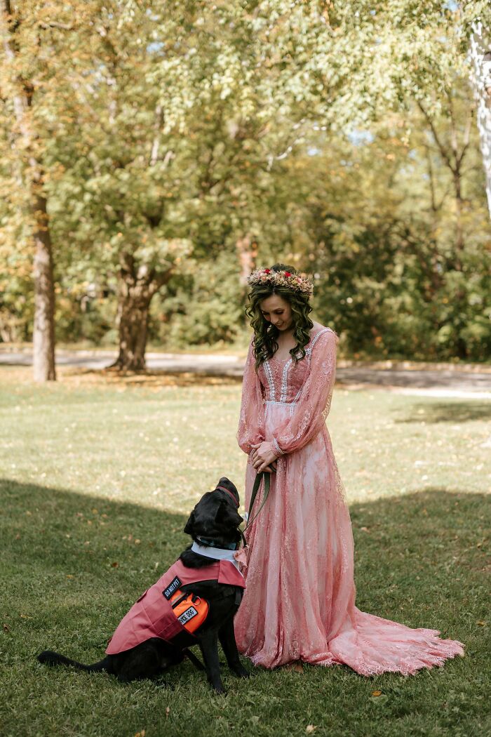 Woman in a pink lace dress with floral crown standing outdoors next to a black dog wearing a service vest, cottage core aesthetics.