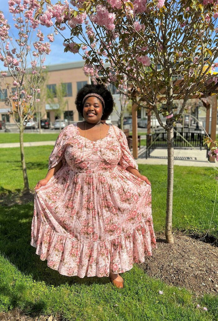 Smiling woman in a floral dress stands under blooming pink trees, showcasing cottage core aesthetics in a sunny outdoor setting.